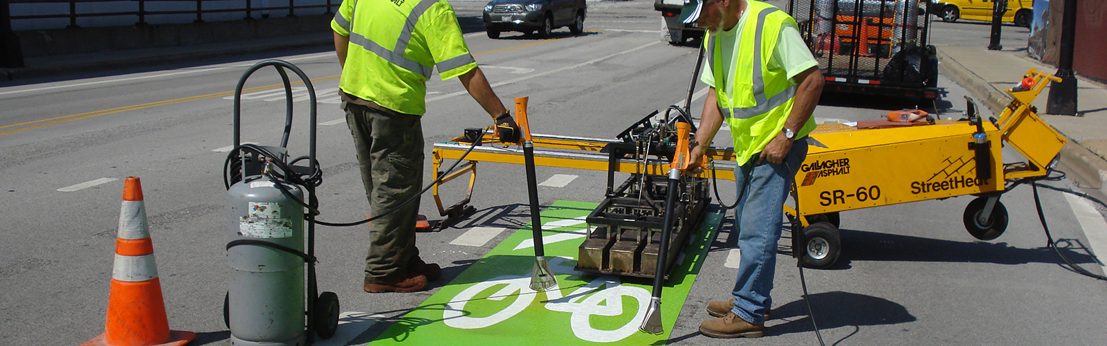 Construction workers in street with construction equipment surrounding as they install green mike symbol marking.