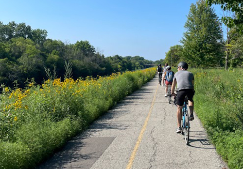 Line of people biking away from viewer on a trail with trees and flowers surrounding