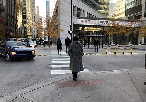 Person crossing street with rubber speed bump and vertical delineators along street centerline and left turning car pausing to yield to pedestrian