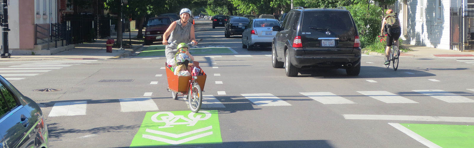 Adult riding cargo bike with children in it and green bikeway markings visible