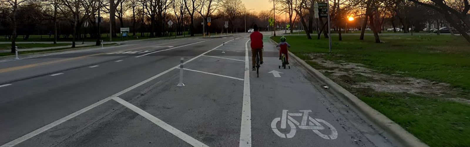 Adult and child riding bikes in bike lane protected from vehicle travel lane by plastic bollards with sunset in background