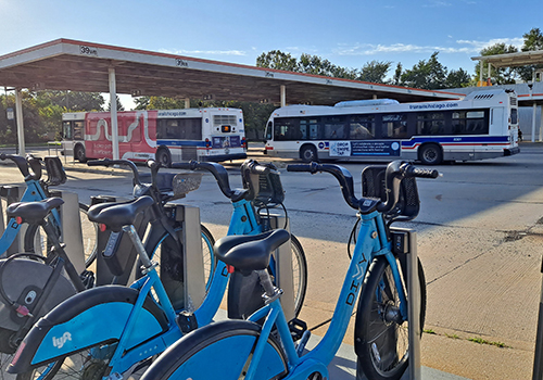 Divvy bikeshare station with a line of bikes in foreground with two CTA buses ined up at transit station in background