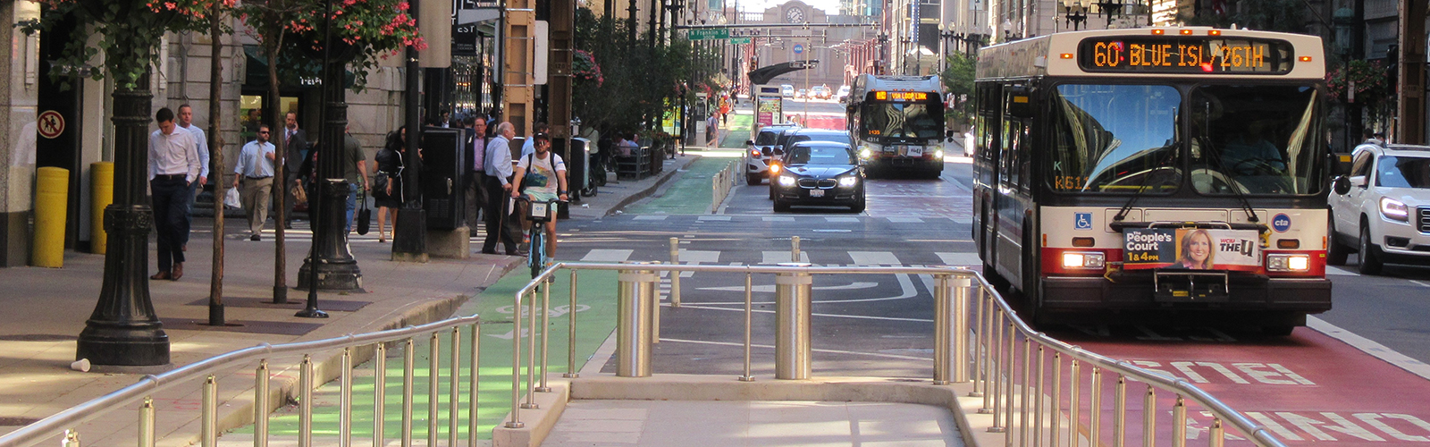 View of dense downtown roadway with oncoming CTA bus separated by bus boarding island and green bike lane with a cyclist biking towards the viewer.