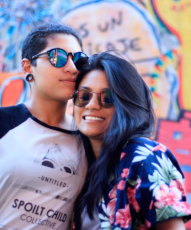 Photo of two women in sunglasses smiling and leaning against each other in front of graffiti wall