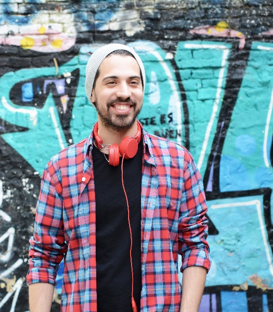 A young bearded male wearing stocking cap, flannel, and red headphones, standing against graffiti wall