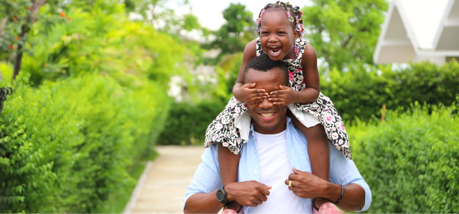 A father with his daughter on his shoulders, covering his eyes with her hands and laughing